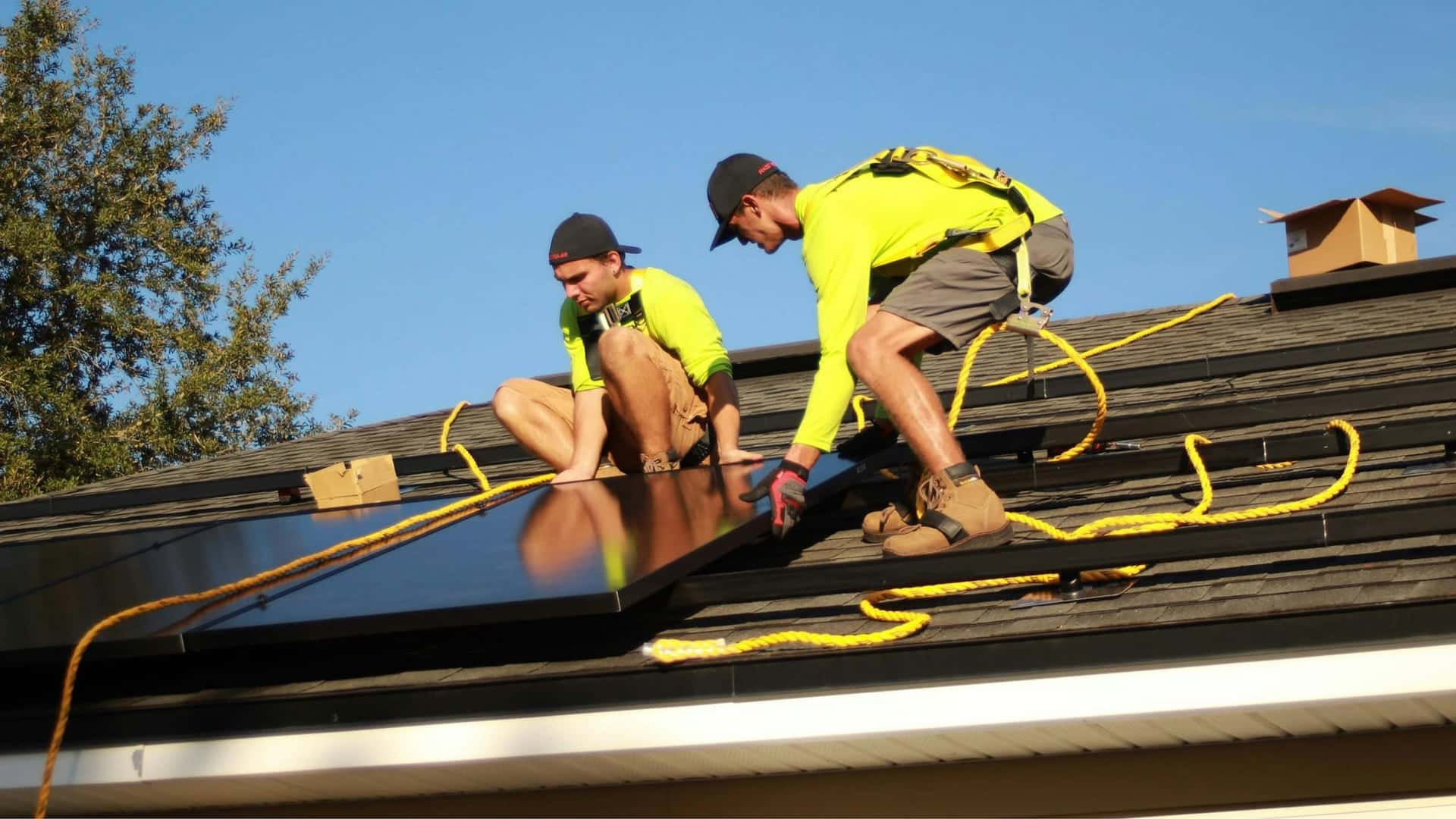 Two solar installers securing panels on a composition-shingle roof under a clear sky